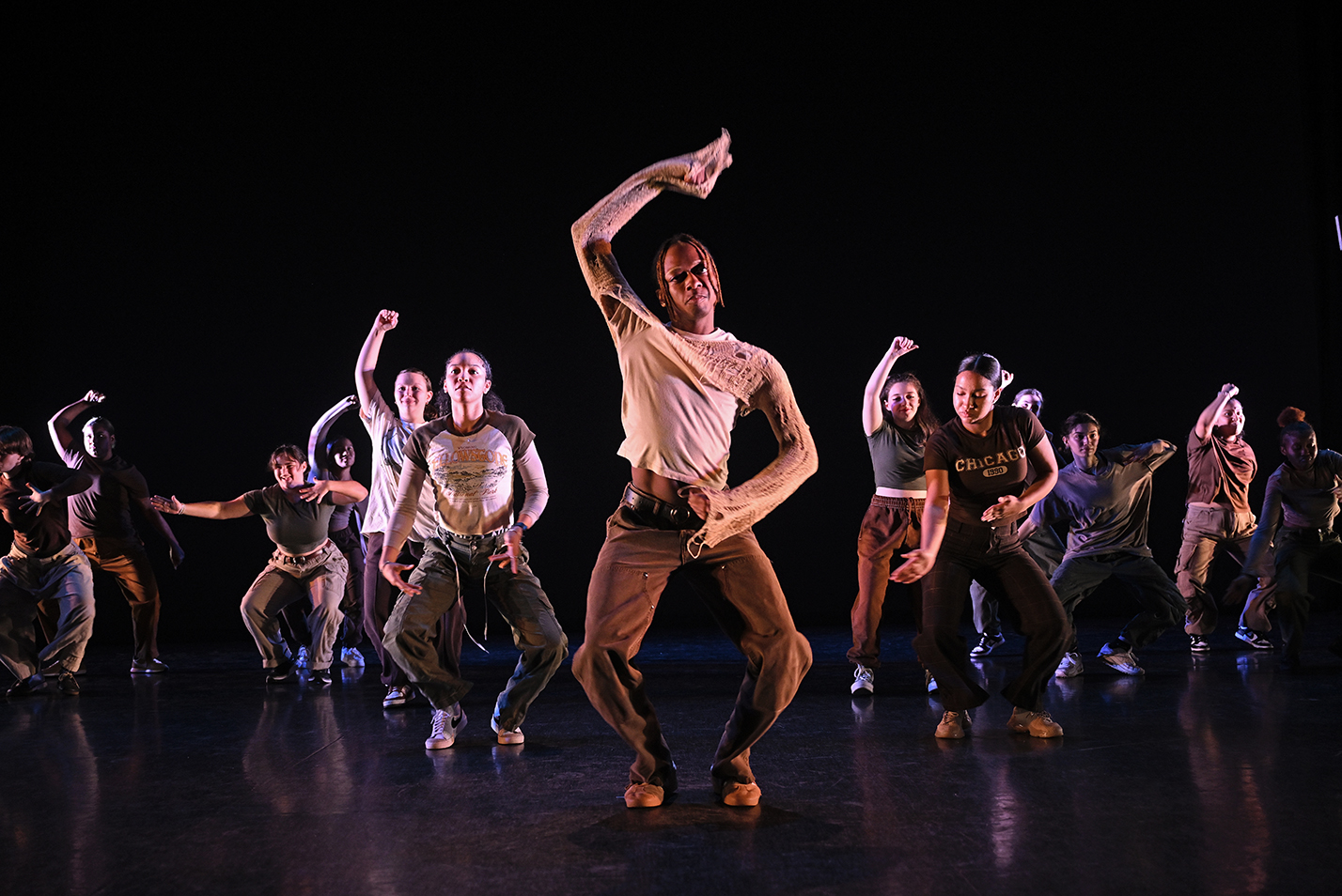 A large group of dancers, fading back from one in the front and center, perform a hip hop dance on stage.